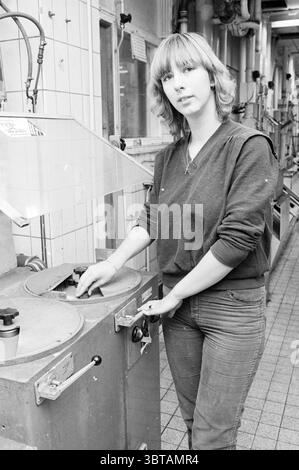 First female process operator Hoogovens Hoogovens, Whizgle News, Dutch Desk, The Netherlands, 1950 - 2000 on 12-11-1981. These topics are shown in the image. The scene depicts a young woman standing in an industrial setting, characterized by a combination of metal machinery and tiled surfaces. She has shoulder-length, wavy hair and appears to be focused, with a serious expression on her face, suggesting concentration or deep thought. The woman is positioned next to a large, circular machine with dials and levers, which she is interacting with by placing her hands on a control. She is dressed i Stock Photo