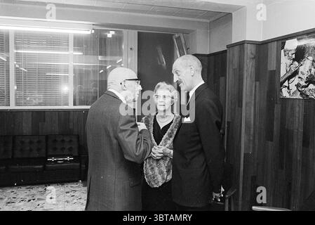Reception Mr. van Vliet Receptions, Whizgle News, Dutch Desk, The Netherlands, 1950 - 2000 on 13-01-1968. These are the elements in the image. The scene captures a moment inside a room characterized by its retro decor and warm, muted tones. Three elderly individuals engage in conversation, forming a small gathering that conveys a sense of intimacy and nostalgia. The man on the left, dressed in a suit with a patterned tie, has a thoughtful expression as he leans slightly forward, gesturing with his hands. His glasses add to his scholarly appearance, drawing attention to his expressive face. Nex Stock Photo