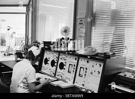Cardiac care department of the Zeeweg hospital Hospitals IJmuiden Zeeweg The Netherlands, Whizgle News, Dutch Desk, The Netherlands, 1950 - 2000 on 22-08-1974. The image shows these topics. The scene captures a vintage control room with a focus on a nurse seated at a large, complex set of equipment. The nurse is dressed in a traditional white uniform and a cap, her attention directed towards a panel filled with dials, buttons, and screens, suggesting a medical or technical environment. The colors are predominantly muted, with shades of gray, white, and black, creating a somewhat nostalgic and Stock Photo