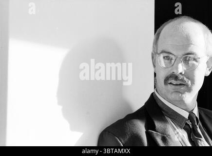 Toon Gerbrands volleyball coach, Whizgle News, Dutch Desk, The Netherlands, 1950 - 2000 on 10-10-1994. These are the elements in the image. In the scene, a middle-aged man with a neatly trimmed mustache and glasses stands slightly off-center, positioned against a stark, light-colored wall. He wears a dark blazer over a collared shirt, which features subtle stripes. The contrast between his attire and the background creates a striking visual dynamic. The lighting is soft yet dramatic, with a beam of light casting a strong shadow on the wall beside him. This shadow adds depth to the composition Stock Photo