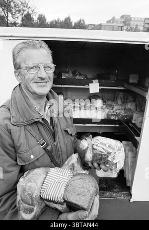 Bakker van Bakel Haarlem The Netherlands, Whizgle News, Dutch Desk, The Netherlands, 1950 - 2000 on 08-09-1993. These topics appear in the image. The scene features an elderly man standing in front of an open display filled with various types of bread. He has silver-gray hair and wears glasses that sit just above his nose. His expression is friendly, characterized by a warm smile that suggests a sense of pride in his offerings. He is dressed in a rugged, dark jacket, hinting at a practical, well-worn style suited for outdoor environments. In his hands, he holds two types of bread: one is a rou Stock Photo