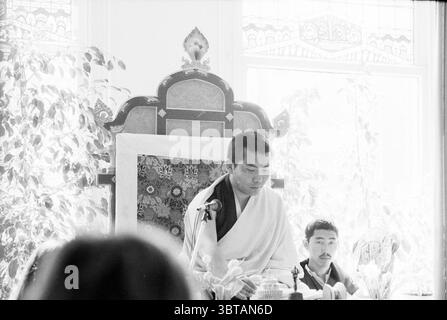 Tibetan Buddhism Haarlem The Netherlands, Whizgle News, Dutch Desk, The Netherlands, 1950 - 2000 on 10-04-1991. The image includes these topics. In a bright and serene setting, a spiritual leader attired in traditional robes stands in front of a beautifully adorned throne. The robes are predominantly white, reflecting a sense of purity and tranquility. The intricate design of the throne, with its dark wood and colorful fabric, adds an element of elegance to the scene. Surrounding this central figure are soft hints of greenery from potted plants, creating a calming atmosphere. Sunlight filters Stock Photo