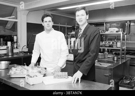 Chef Greveling and Peter Bruins Bokkedoorns Restaurant, Whizgle News, Dutch Desk, The Netherlands, 1950 - 2000 on 10-01-1989. The image contains these topics. In a bustling kitchen, two individuals stand together at a stainless steel worktable. The first person, dressed in a crisp white chef's uniform, appears focused and confident. His short, dark hair is neatly styled, and he looks directly at the observer with a friendly expression. He stands beside the second individual, who is clad in a dark, stylish suit and tie, exuding a polished and professional demeanor. The kitchen is filled with an Stock Photo