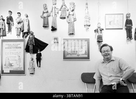 Man at puppet exhibition, Whizgle News, Dutch Desk, The Netherlands, 1950 - 2000 on 04-2000. The image shows these topics. The scene captures a cozy, artistic space dedicated to puppetry. A range of intricately crafted puppets hangs against a light-colored wall, showcasing various expressions and styles, from whimsical to more traditional figures. The puppets are suspended by thin strings and vary in size, featuring vivid details like painted faces and ornate costumes. To the left, an older man sits casually on a dark chair, wearing spectacles and a light-colored shirt, which adds a soft contr Stock Photo