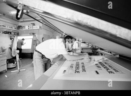Screen printers at work, Whizgle News, Dutch Desk, The Netherlands, 1950 - 2000 on 19-05-1992. The image includes these topics. The scene depicts a busy workspace filled with a mix of industrial and artistic elements. In the foreground, a person is focused intently on the task at hand, leaning forward with one hand resting on a large sheet laid out on a flat surface. The individual is dressed in a simple, light-colored shirt, and has a concentrated expression, suggesting dedication to their work. The workspace is well-lit, with overhead fluorescent lights casting a bright, even glow. This illu Stock Photo