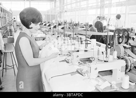 Woman near a row of sewing machines., Whizgle News, Dutch Desk, The Netherlands, 1950 - 2000. These topics are shown in the image. In a bustling workshop setting, a woman with a stylish bob haircut is engaged in her work at a sewing machine, exuding concentration and dedication. She wears a fitted, sleeveless dress that enhances her figure, adding a touch of elegance amidst the industrial environment. The sewing machine in front of her is surrounded by spools of thread in various colors, showcasing a range of vibrant hues, although the scene is primarily rendered in monochrome, lending a timel Stock Photo