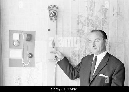 Mr. Ramakers Alarm Institute Persons, Whizgle News, Dutch Desk, The Netherlands, 1950 - 2000 on 21-08-1967. These topics appear in the image. The scene is set in a room characterized by a vintage atmosphere. In the foreground stands a man dressed in a sharply tailored suit. His hairstyle is neatly combed, and he appears poised and confident as he gestures towards a wall-mounted clock positioned on a wooden board. The clock has distinct features, with a classic round face showcasing bold numbers, marking the time with elegant hands. Surrounding the clock, there are various objects, including a Stock Photo