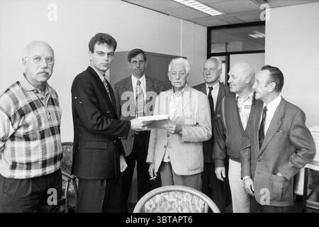 Handing over signatures to Alderman Pont Hoofddorp The Netherlands, Whizgle News, Dutch Desk, The Netherlands, 1950 - 2000 on 16-09-1996. These are the topics in the image. The scene captures a group of seven individuals in a modest, well-lit indoor setting. The space has a professional ambiance, characterized by neutral-colored walls and bright overhead lighting that casts a clean, even glow. In the foreground, a young man stands slightly off-center, wearing a dark suit and a tie. He is engaging with an older gentleman, who appears to be receiving a document. The older man, with white hair an Stock Photo