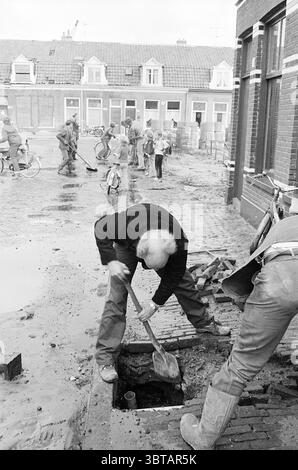Hole in the street through water Barendsestraat Streets Water, Whizgle News, Dutch Desk, The Netherlands, 1950 - 2000 on 10-09-1976. These topics are shown in the image. The scene captures a moment of community effort in a gritty, urban environment. Several individuals are engaged in physical labor, working together on what appears to be a street construction or repair task. In the foreground, an elderly man with white hair bends over a deep, dark hole, wielding a shovel with determination. He is dressed in practical, rugged clothing suited for labor, and his concentration is evident as he dig Stock Photo