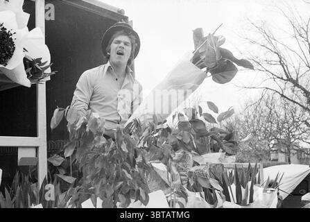 Seller of flowers and plants on the street, Whizgle News, Dutch Desk, The Netherlands, 1950 - 2000 on 27-04-1979. The image includes these topics. In a bustling market scene, a young man stands at the back of a truck, which is partially filled with a vibrant array of potted plants and fresh flowers, their colors contrasting against a muted background. The man, wearing a wide-brimmed hat, appears animated, possibly calling out to passersby or expressing excitement. His expression is lively, with his mouth open mid-sentence. The flowers around him showcase an assortment of greens, pinks, and yel Stock Photo
