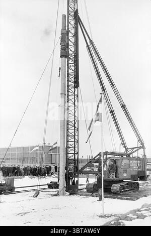 1st pile workshop the Tunnel First pile, Whizgle News, Dutch Desk, The Netherlands, 1950 - 2000 on 05-03-1971. These topics are shown in the image. In a wintry outdoor setting, a tall drilling rig dominates the scene, its metallic structure rising prominently against the lightly overcast sky. The rig is an industrial gray, contrasting sharply with the surrounding blanket of white snow. Its upper portion is intricately designed for operation, featuring cables and pulleys that hang down, adding to the complex visual texture of the structure. Nearby, large machinery, specifically a crawler crane, Stock Photo