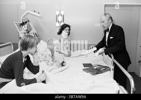 Magic in the Deodorant for patients Magic, Whizgle News, Dutch Desk, The Netherlands, 1950 - 2000 on 16-02-1981. These topics are shown in the image. In a bright, sterile room, a hospital bed dominates the scene, dressed in crisp white sheets. The bed is surrounded by three individuals: two children, sitting close together, and an adult standing beside them. The children, a boy and a girl, have a subdued demeanor, reflecting their surroundings. The boy is dressed casually, while the girl wears a patterned top, her hair adorned with a ribbon. The adult, distinguished in appearance with silver h Stock Photo