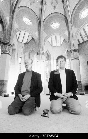 Quiet room Bloemendaal Miscellaneous Interior Bloemendaal, Whizgle News, Dutch Desk, The Netherlands, 1950 - 2000 on 15-02-1989. These are the elements in the image. The scene captures two individuals engaged in meditation within a spacious, serene interior. They are positioned on their knees on a soft, neutral-toned surface, which creates a calming effect. The architecture around them features tall, arched ceilings with intricate brickwork, giving a sense of grandeur and peace. Natural light pours in through large stained-glass windows, casting colorful patterns and subtle hues across the sto Stock Photo