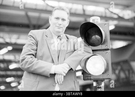 Mr. Black referee, Whizgle News, Dutch Desk, The Netherlands, 1950 - 2000 on 16-10-1991. The image contains these topics. The scene features a man standing relaxed next to a traffic signal. He is dressed in a tailored suit that contrasts with the industrial setting behind him, suggesting a mid-20th century aesthetic. His shirt is likely a lighter color, while the tie adds a touch of formality. The traffic signal stands tall beside him, with its familiar red and green lenses prominently visible. The surrounding environment appears to be an indoor space, possibly a transport hub, characterized b Stock Photo