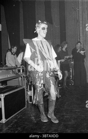 Mendel College fashion show, Whizgle News, Dutch Desk, The Netherlands, 1950 - 2000 on 12-11-1990. The image includes these topics. In a dimly lit venue, a lively scene unfolds on stage, creating an energetic atmosphere. A young individual is at the center, dressed in an unconventional costume made of various materials, including what appears to be strips of fabric in earthy tones. The attire has a playful yet artistic flair, contributing to a vibrant visual dynamic. Surrounding the central figure, several people are engaged in different activities. To the left, a DJ is busy at a soundboard, s Stock Photo