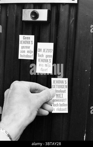 Anke Skelers Jehovah's Witness, Whizgle News, Dutch Desk, The Netherlands, 1950 - 2000 on 16-05-1994. The image shows these topics. The scene features a hand reaching towards a dark, textured surface, which appears to be a door made of metal or a similar material. The surface has a corrugated pattern, creating a sense of depth. A small circular object, likely a doorbell or a small camera, is mounted above the door, contrasting with the rugged background. Three distinct rectangular cards are affixed to the door. These cards are predominantly white with bold, contrasting text in black, drawing a Stock Photo