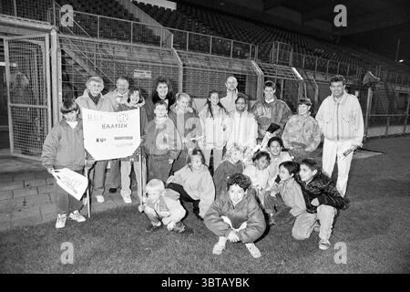 School guest at the Haarlem - NEC match Haarlem The Netherlands, Whizgle News, Dutch Desk, The Netherlands, 1950 - 2000 on 25-10-1991. These topics are shown in the image. The scene unfolds in a large, open space, likely an athletic stadium, characterized by a vast field of green grass that leads to a backdrop of bleachers. The bleachers are empty, with shadows indicating the absence of spectators and lending a slightly somber atmosphere to the setting. Centered in the lower part of the scene, a diverse group of individuals gathers closely together, displaying a mix of ages and emotions. Among Stock Photo