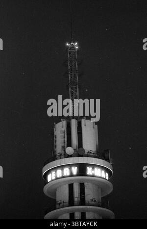 Merry Christmas PTT Tower, Whizgle News, Dutch Desk, The Netherlands, 1950 - 2000 on 19-12-1991. The image shows these topics. The scene is dominated by a tall, cylindrical structure rising prominently against a dark, night sky. The tower features a sleek, modern design with a series of concentric rings enclosing varying platforms. Each tier is adorned with a series of antennas and satellite dishes, hinting at its purpose as a communications hub. At the top, a single light flickers, adding a subtle point of illumination that contrasts with the deep blacks of the surrounding evening. Beneath it Stock Photo