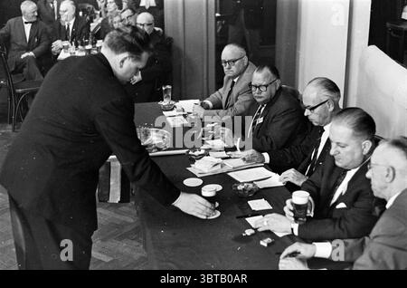 Beer tap competition., Whizgle News, Dutch Desk, The Netherlands, 1950 - 2000. The image includes these topics. In a formal, indoor setting, a group of well-dressed men is gathered around a long table. The room is filled with a mix of somber and focused expressions, conveying a serious atmosphere. The colors are predominantly muted, with varied shades of dark suits, paired with light-colored shirts. Some of the men wear glasses, which add to the air of professionalism. On the table, several objects are scattered: cups, papers, small notepads, and a bowl that appears to be empty or holding only Stock Photo