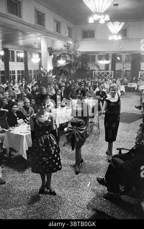 Models from Institut d'Elegance show fashion from fashion house Léon in Dreefzicht Haarlem. Fashion show. Haarlem The Netherlands, Whizgle News, Dutch Desk, The Netherlands, 1950 - 2000 on 30-10-1960. The image includes these topics. The scene captures a vibrant indoor setting, reminiscent of a mid-20th-century event. The room is spacious and filled with round tables draped in white tablecloths, where elegantly dressed guests sit in clusters, engaged in conversation. Three models elegantly walk along a runway that extends into the dining area, each showcasing a distinct style. The first model Stock Photo