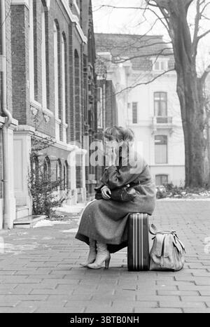 Margot room occupant sitting on suitcase Haarlem Parklaan The Netherlands, Whizgle News, Dutch Desk, The Netherlands, 1950 - 2000 on 22-02-1979. These are the topics in the image. The scene unfolds in a monochromatic palette, conveying a sense of nostalgia and quiet introspection. A young woman, dressed in a long, tailored coat, sits on a sturdy suitcase positioned on the cobblestone pavement. Her posture is a mix of contemplation and waiting, as she rests her chin on her hand, gazing off into the distance. She has a subtle air of elegance, accentuated by knee-high boots, which lend a stylish Stock Photo