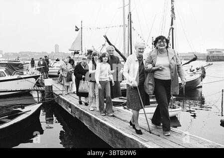 Senior Citizens' Day at IJmuiden Marina Ships, Whizgle News, Dutch Desk, The Netherlands, 1950 - 2000 on 14-09-1974. The image includes these topics. In a bustling harbor scene, several people are gathered on a wooden dock that extends over calm waters. The setting is filled with boats of varying sizes, some gently bobbing on the water, reflecting the overcast sky. Toward the foreground, a group of individuals walks along the dock. They are a mix of ages, with a prominent elderly woman using a cane for support, her silver hair contrasting with the darker clothing of those around her. Next to h Stock Photo