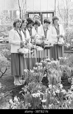 Keukenhof on opening day flower girls in greenhouse Lisse, Whizgle News, Dutch Desk, The Netherlands, 1950 - 2000 on 22-03-1978. These topics appear in the image. In a serene garden setting filled with blooming flowers, a group of women stands together, each holding a wicker basket filled with an arrangement of blossoms. They are dressed in coordinated outfits featuring light-colored blazers and knee-length skirts, creating a unified, elegant look. The fabric is soft, likely in pastel hues that complement the vibrant colors of the surrounding flora. The women exhibit a variety of hairstyles, w Stock Photo