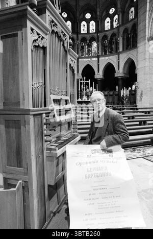 Organist Bartelink in Basilica, Whizgle News, Dutch Desk, The Netherlands, 1950 - 2000 on 16-12-1990. These are the elements in the image. The scene captures an interior of a grand, Gothic-style cathedral characterized by soaring arches and intricate stained glass windows. Sunlight filters through the colorful glass, casting a kaleidoscope of hues onto the stone floor, which creates a rich contrast of light and shadow. In the foreground, an older man sits beside a large organ, meticulously crafted from dark wood with ornate details that highlight its significance in the space. He has a thought Stock Photo