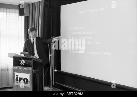 IRO meeting at Radio Holland Amsterdam Meetings Amsterdam The Netherlands, Whizgle News, Dutch Desk, The Netherlands, 1950 - 2000 on 31-05-1988. These topics are shown in the image. The scene features a well-dressed man standing in front of a podium, gesturing emphatically as he delivers a presentation. He has dark hair and is wearing glasses, giving him a scholarly appearance. The setting exudes a professional atmosphere, possibly within a conference room or seminar hall. In the background, a large screen displays a list of topics, arranged in bullet points. The lighting is a mix of natural l Stock Photo