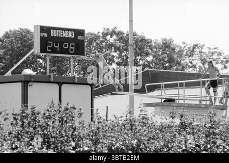 Groenendaal swimming pool Heemstede The Netherlands, Whizgle News, Dutch Desk, The Netherlands, 1950 - 2000 on 19-07-1990. The image shows these topics. The scene captures an outdoor swimming pool environment under bright, clear skies, suggesting a warm, sunny day. The focus is on a diving platform from which a person is poised to leap into the water below. This individual, dressed in light swim trunks, is captured mid-action, illustrating a sense of excitement and anticipation as they prepare to dive. In the background, another swimmer stands near the edge of the platform, watching the diver Stock Photo