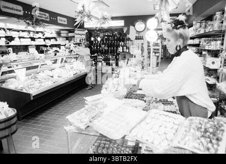Interior of wine and delicatessen store Leo Zwarter Heemstede Heemstede The Netherlands, Whizgle News, Dutch Desk, The Netherlands, 1950 - 2000 on 02-03-1989. These topics appear in the image. In a vibrant deli shop filled with an array of items, the scene is bustling with activity. To the left, a showcase displays a colorful assortment of cheeses, with various shapes and textures. Each cheese is neatly arranged, showcasing a spectrum of yellows and whites, some with distinct dark rinds. In the foreground, a woman with a stylish haircut, dressed in a chunky, oversized sweater, is carefully arr Stock Photo