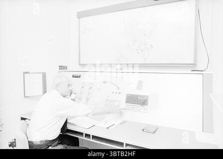 Commissioning of connection equipment for the Beverwijk fire brigade. Fires fire brigade Commissioning commissioning, Whizgle News, Dutch Desk, The Netherlands, 1950 - 2000 on 26-08-1977. The image includes these topics. The scene features a man sitting at a control station, characterized by a clean, white environment that exudes a sense of simplicity and focus. He is dressed in a formal white shirt, looking intently at a series of controls and screens before him. The control panel is fairly minimalist, adorned with various dials, switches, and a large monitor displaying a map of a region, sug Stock Photo