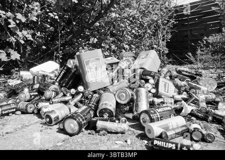 Mountain of old batteries, Whizgle News, Dutch Desk, The Netherlands, 1950 - 2000 on 21-04-1991. These are the elements in the image. The scene depicts a chaotic scattering of discarded batteries in a natural setting. Various brands, notably Philips and others, are discernible among the heap, each one showing its distinctive colors and designs, though muted due to the monochromatic presentation. The batteries, in different sizes and shapes, intersperse with some larger rectangular battery packs. Surrounding the litter are hints of green foliage, indicating that this area might be on the edge o Stock Photo