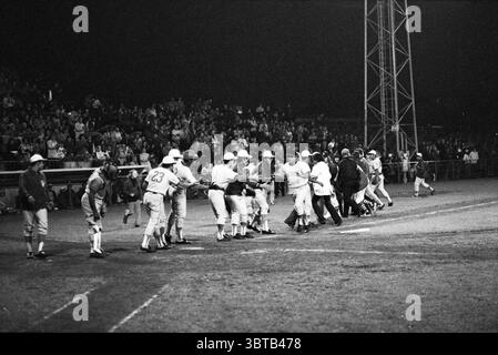 Cuba - Nicaraqua Baseball, Whizgle News, Dutch Desk, The Netherlands, 1950 - 2000 on 30-06-1972. These are the elements in the image. The scene captures a lively baseball game under the glow of bright stadium lights, illuminating the field and creating dramatic contrasts in the dark surroundings. Players in light-colored uniforms, featuring a mix of white and light blue, are actively engaged in handshakes and high-fives, indicating a celebratory moment, perhaps after a significant win or play. The crowd is composed of numerous spectators, their silhouettes outlined against the light, showcasin Stock Photo