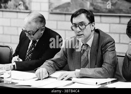 C.O.R. meeting VNU Hoog Brabant Utrecht VNU Association of Dutch Publishers Utrecht, Whizgle News, Dutch Desk, The Netherlands, 1950 - 2000 on 27-11-1981. These are the elements in the image. In a conference room setting, two men are seated at a rectangular table. The man in the foreground is engaged in discussion, wearing dark-rimmed glasses and a gray plaid suit over a white shirt and patterned tie. His expression is serious, and he gestures with one hand while the other rests on papers spread before him. The background features a textured wall, possibly made of light-colored bricks, providi Stock Photo