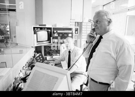 Two doormen at the Spaarne Hospital, Whizgle News, Dutch Desk, The Netherlands, 1950 - 2000 on 30-04-1995. These topics appear in the image. The scene captures two individuals within a control room setting, characterized by a blend of technology and a sense of focused activity. The first person, seated, has an intent expression, with a computer monitor displaying data in front of him. He is dressed in a light-colored shirt, and his attire conveys a formal yet approachable air. The second individual stands nearby, engaged in a conversation over the phone, holding the receiver to his ear. He is Stock Photo