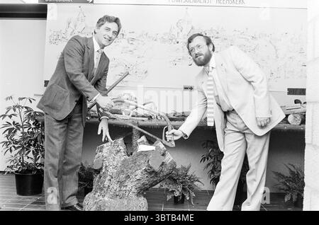 25 years Avg. works Heemskerk Jubilee anniversaries Heemskerk, Whizgle News, Dutch Desk, The Netherlands, 1950 - 2000 on 06-05-1982. These are the topics in the image. The scene features two men standing beside a large, rough-hewn wooden stump that serves as a focal point. Both individuals are engaged in a shared activity, each holding a tool that appears to be a saw, poised as if to cut the stump. The man on the left is dressed in a light-colored suit with a patterned tie, exuding a sense of formality and professionalism. His posture suggests enthusiasm and engagement in the task. The man on Stock Photo