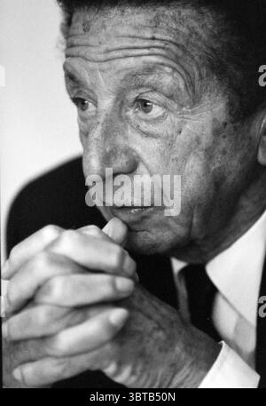 Mr. Orlandini ex KLM, Whizgle News, Dutch Desk, The Netherlands, 1950 - 2000 on 11-10-1993. The image contains these topics. The scene presents an elderly gentleman, whose face is a canvas of time, displaying deep lines and a textured surface that speaks of experience. His hair is dark, possibly graying, and neatly styled, contributing to his composed demeanor. He rests his chin on his hand, fingers interlocked, which reveals slender, aged hands with visible veins. The elegance of his attire—a dark suit—contrasts with the softness of his skin. The subdued palette of blacks and grays evokes a c Stock Photo