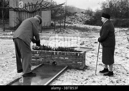 Two elderly people playing on a miniature golf course., Whizgle News, Dutch Desk, The Netherlands, 1950 - 2000. These are the elements in the image. In a somber landscape, a man and an elderly woman are engaged in a quiet moment. The ground is covered with a light dusting of snow, giving the scene a stark, wintry appearance. The man, dressed in a heavy, winter coat and a hat, is bent over a low stone structure, tending to what appears to be a small garden or patch of plants. His concentration is evident as he carefully examines the greenery, contrasting against the monochrome environment. The Stock Photo