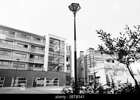 A flat., Whizgle News, Dutch Desk, The Netherlands, 1950 - 2000. The image shows these topics. The scene features a modern residential area characterized by sleek architecture. The setting consists of several buildings, predominantly constructed with a combination of concrete and glass, showcasing large windows that reflect the sky. The color palette is primarily monochromatic, with varying shades of gray and white dominating the structures, giving a clean, contemporary feel. A slim, elegant streetlamp stands prominently at the forefront, its lantern casting a geometric shadow on the ground. S Stock Photo