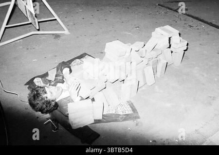 Laughing man under a 'blanket' of stones in front of café 't Haasje Haarlem The Netherlands, Whizgle News, Dutch Desk, The Netherlands, 1950 - 2000 on 18-05-1981. These are the topics in the image. The scene depicts a young individual lying on a patterned blanket spread across a hard, possibly concrete, floor. They appear to be positioned in a relaxed or slumberous state, with their body partially obscured by a stack of rectangular, unadorned bricks arranged haphazardly around them. The bricks have a muted, earthy tone, blending subtly into the surrounding environment. The lighting is somewhat Stock Photo