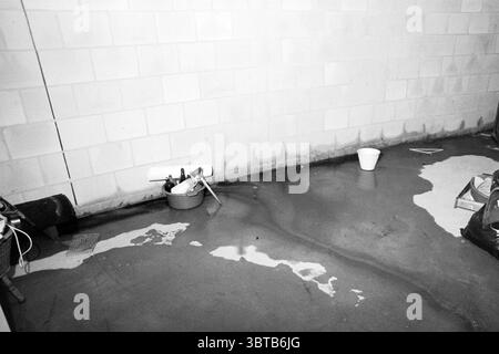 The new construction of the Overduin apartment complex on the Domploodslaan in Bloemendaal is suffering from excessive water. Bloemendaal Dompvloedslaan, Whizgle News, Dutch Desk, The Netherlands, 1950 - 2000 on 30-10-1994. These topics appear in the image. The scene depicts a dimly lit basement or storage area with pale, unfinished cement walls. The floor is primarily smooth and shiny, reflecting dim light, but also shows signs of dampness with scattered puddles of water creating a mottled effect. In one corner, a plastic bucket stands upright, its presence contrasting with the more rugged, u Stock Photo