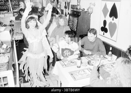 Belly dancer in restaurant Passport Dancing, Whizgle News, Dutch Desk, The Netherlands, 1950 - 2000 on 14-11-1988. The image shows these topics. In a lively indoor setting, a vibrant atmosphere is created by a gathering of people engaged in various activities. The space is characterized by cozy, dim lighting that enhances an intimate mood. Central to the scene is a performer, dressed in a striking costume adorned with feathers and sequins, exuding a sense of celebration and energy as she dances. Her pose, arms raised high, reflects joy and enthusiasm. The colors of her attire—bright and bold—c Stock Photo
