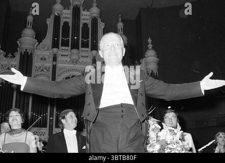 Farewell conductor Katlewitz N.P.O. Conductors, Whizgle News, Dutch Desk, The Netherlands, 1950 - 2000 on 25-04-1982. The image includes these topics. In the scene, a man stands confidently at the center, arms outstretched, conveying a sense of triumph or celebration. He is dressed in a classic tuxedo, featuring a black jacket with a white shirt, suggesting a formal occasion. His expression is joyful, radiating enthusiasm and accomplishment. Surrounding him are several individuals, also dressed in formal attire, reflecting a similar festive atmosphere. The people vary in expression, some smili Stock Photo