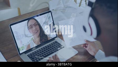 Colleague wearing headphones calling on laptop reviewing printed diagram at office desk, copy space Stock Photo