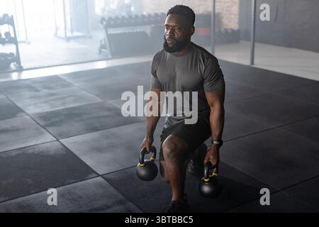 Fit African American man performing kettlebell lunges on gym floor in sportswear near dumbbell rack Stock Photo