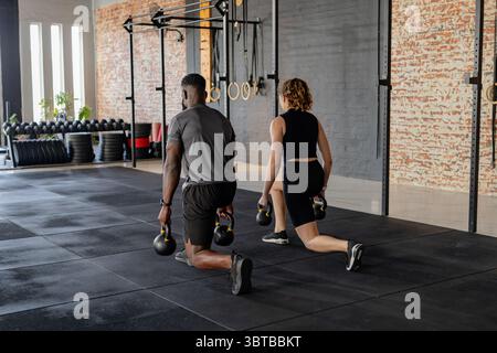 Diverse workout partners in athletic wear doing weighted lunges using kettlebells in CrossFit gym Stock Photo