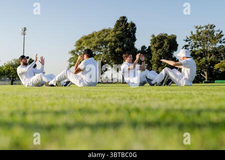 Diverse male teammates doing sit-ups and high-fiving on grass in cricket uniforms under floodlight Stock Photo