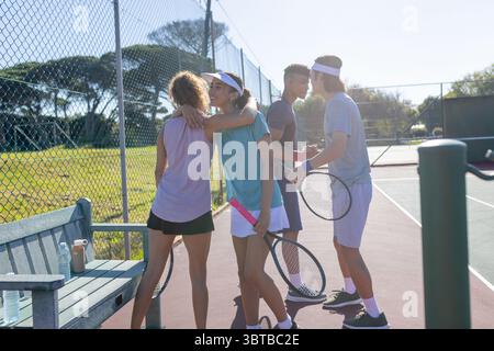 Diverse tennis friends celebrating victory on tennis court holding rackets next to bench Stock Photo