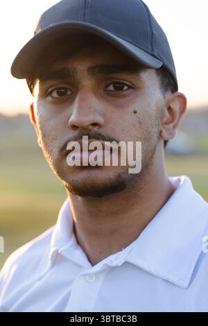 Indian man standing in grassy field during golden hour wearing black baseball cap white polo shirt Stock Photo